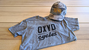Camo Oxyd hat and gray t-shirt on wood table top.