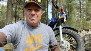 Man wearing Oxyd Cycles Tee in mountains with TW200 motorcycle. 